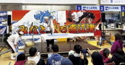 みなとまち直江津と書かれたパネルの前でブリの解体を行う様子(写真)